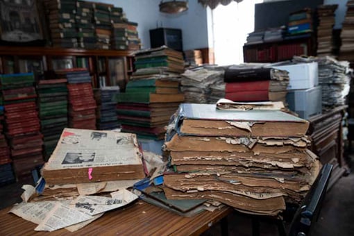 A stack of newspaper volumes on a table at the McMillan Memorial Library in Nairobi, Kenya, with piles of archives on the table and on bookshelves in the background. 