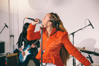 A photograph of a female wearing an orange top singing into a microphone