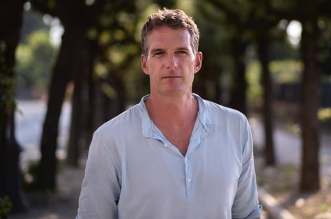 History broadcaster and writer Dan Snow, standing facing the camera in a white shirt, with two rows of trees behind him.  